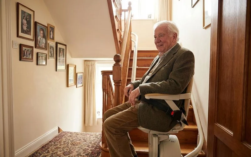Senior man comfortably seated on stair lift with safety belt ascending wooden staircase at home